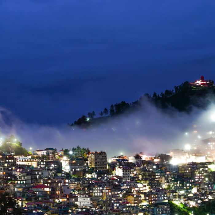 Monsoon mist drifts across the hills as illuminated buildings and houses light up the cityscape, with Jakhu Temple visible atop the hill after rainfall in Shimla
