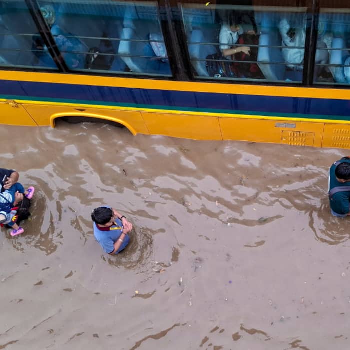 Parents wade through a waterlogged road to drop their children on a school bus following heavy rainfall, at Subhash Chowk in Gurugram, Haryana